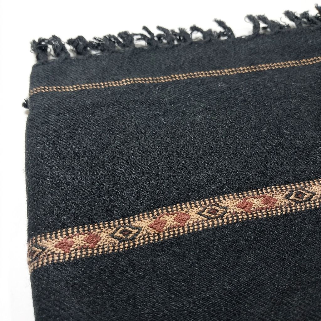 Close-up view of a men's black wool shawl showing woven fabric texture, brown geometric border pattern, and fringe tassels