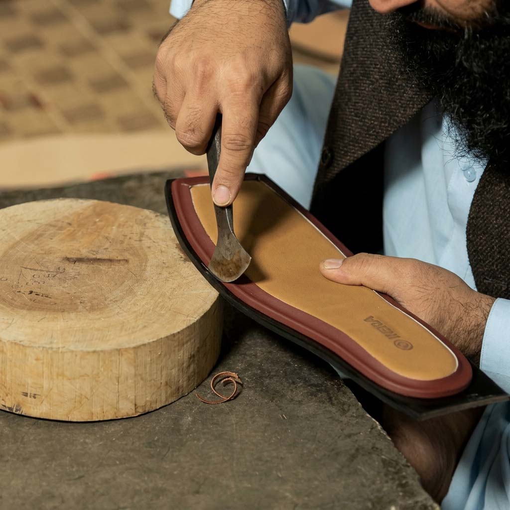 Close-up photograph of a master craftsman in traditional attire meticulously hand-cutting a durable tire rubber outsole for a Bera Peshawari chappal sandal on a wooden block with a traditional Edge Creaser/Skiving Knife.