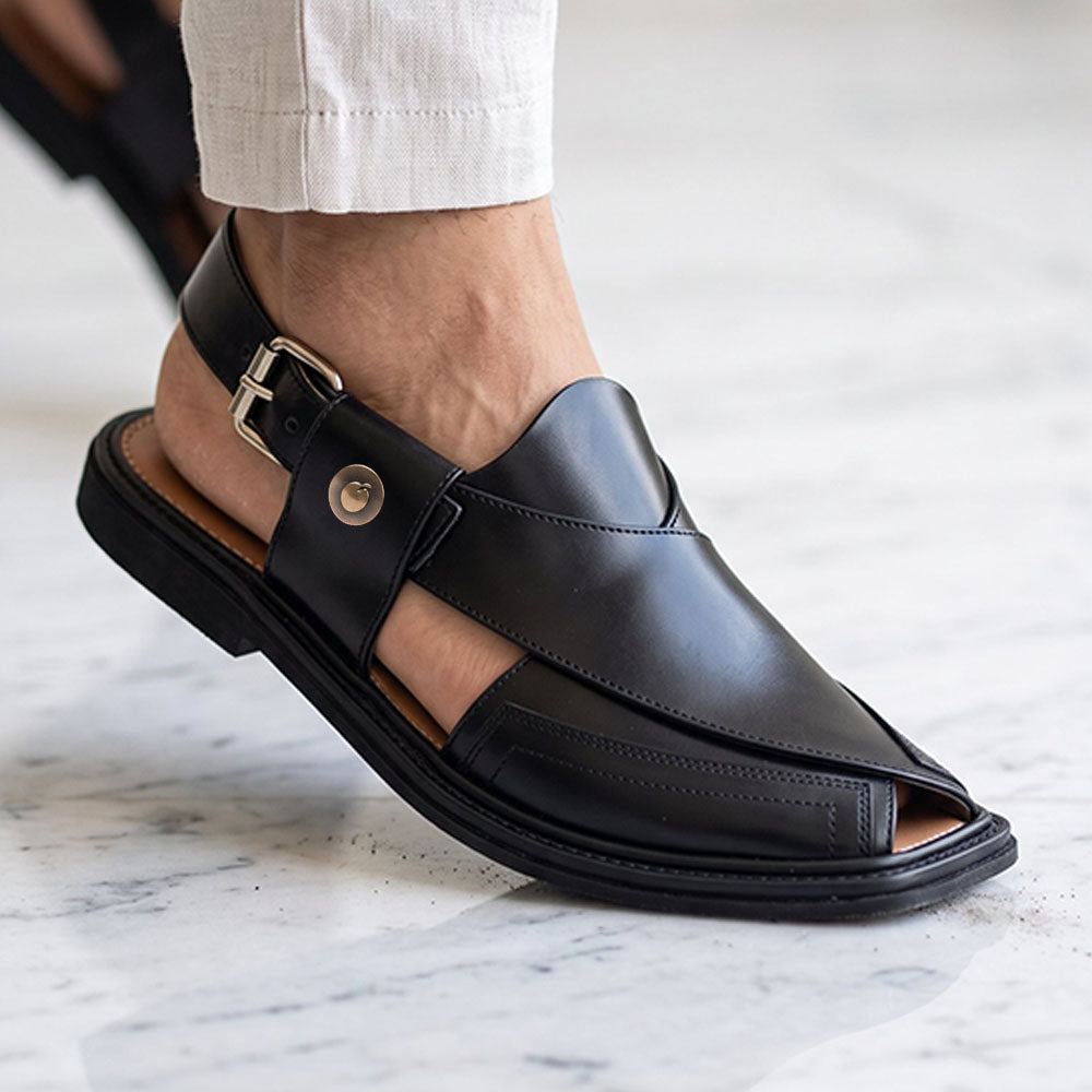 A close-up of a person's foot wearing a Bera black leather open-toe peshawari chappal taki style with a buckle strap and a Bera emblem, paired with light-colored shalwar, standing on a white marble floor.