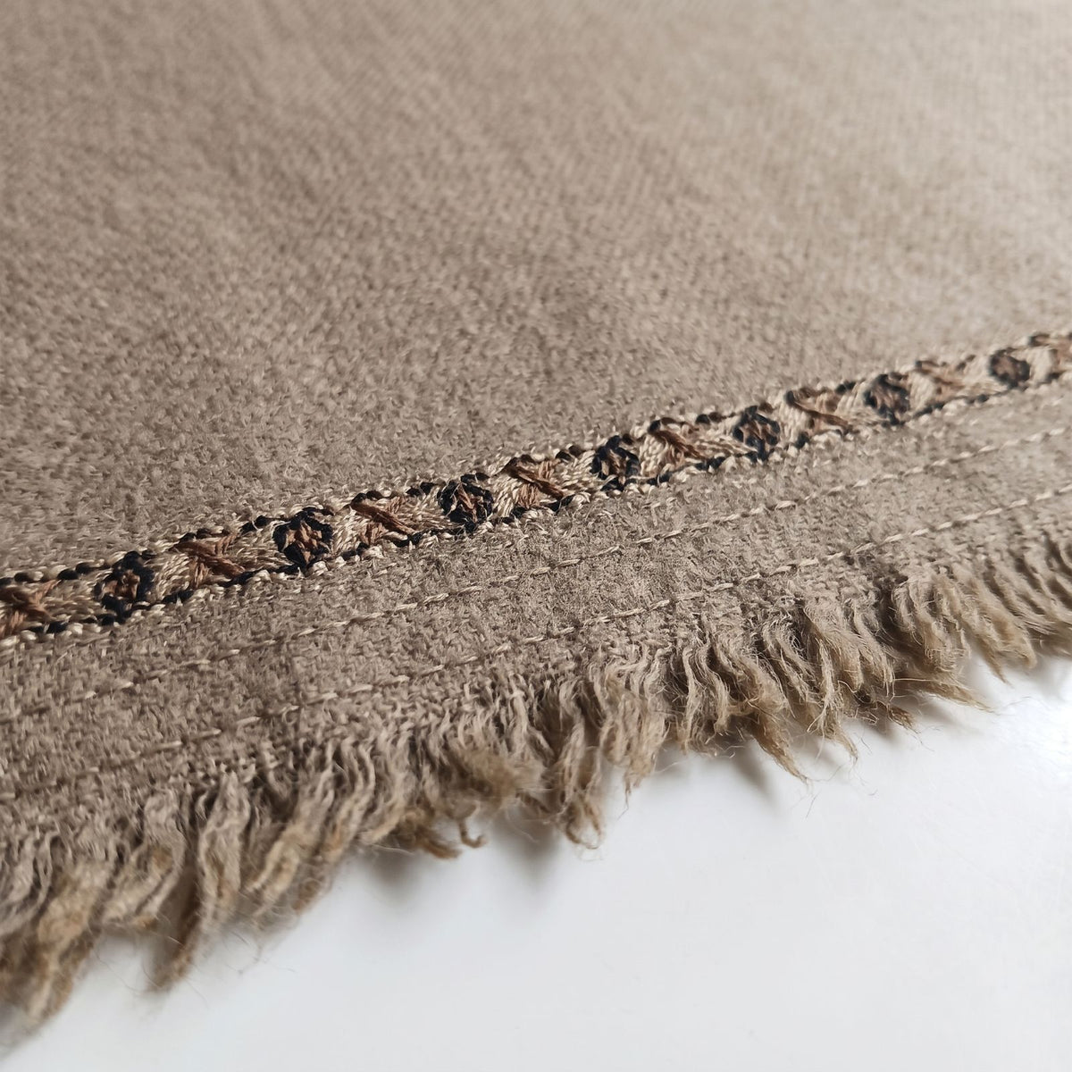 Close-up macro view of a men's beige wool shawl showing textured fabric, woven geometric border, and fringe tassel ends   
