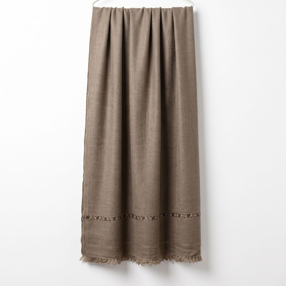 Men's beige wool shawl featuring a traditional woven geometric border and fringe tassels hanging vertically on a display wall   