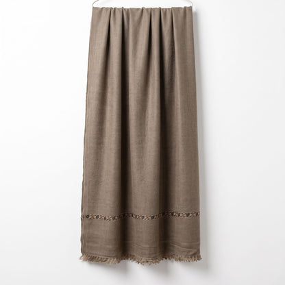 Men's beige wool shawl featuring a traditional woven geometric border and fringe tassels hanging vertically on a display wall   