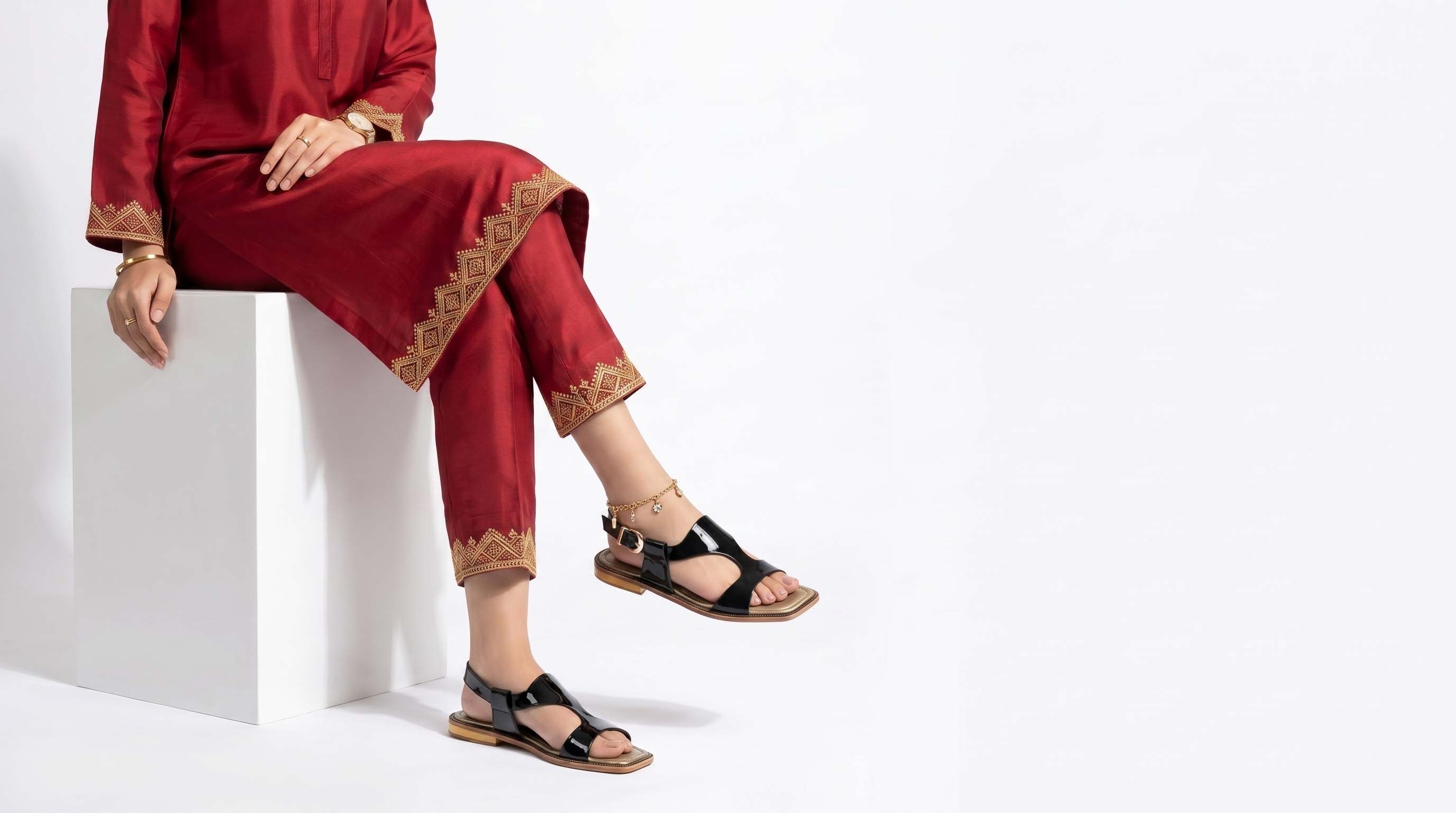 A model wearing a deep red kurta and matching trousers with gold embroidered borders, sitting on a white block. The model is also wearing black ladies peshawari chappal with brown soles.